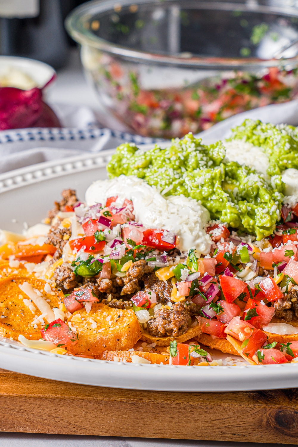 A white platter with sweet potato nachos including sweet potato medallions topped with seasoned ground beef, pico de gallo, cheese, sour cream, and guacamole. The platter is on a wooden board on a marble counter with a blue striped napkin.