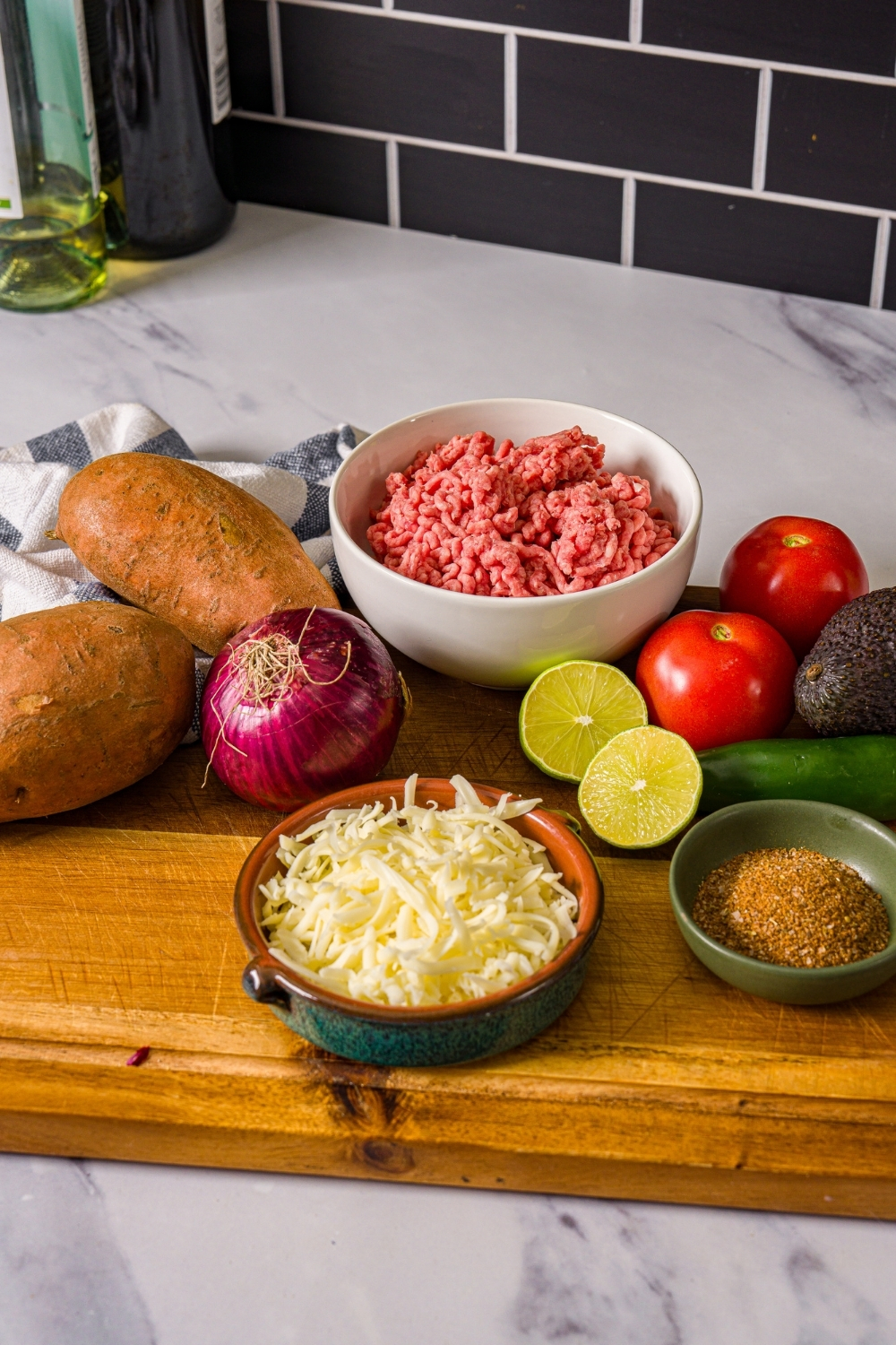 A wooden board with ingredients to make sweet potato taco bowls including ground beef, sweet potatoes, shredded cheese, lime, tomatoes, avocado, red onion, and seasonings.