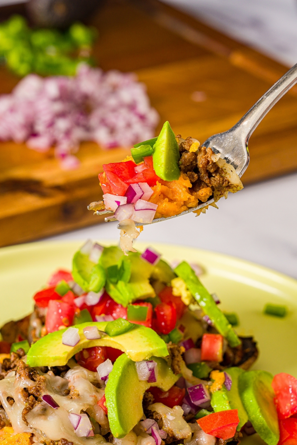 A close up of a fork taking a bite from a plate with a sweet potato taco bowl including seasoned taco meat, avocado, sweet potato, cheese, and chopped tomatoes and red onion. The plate is on a marble counter with a board of toppings.
