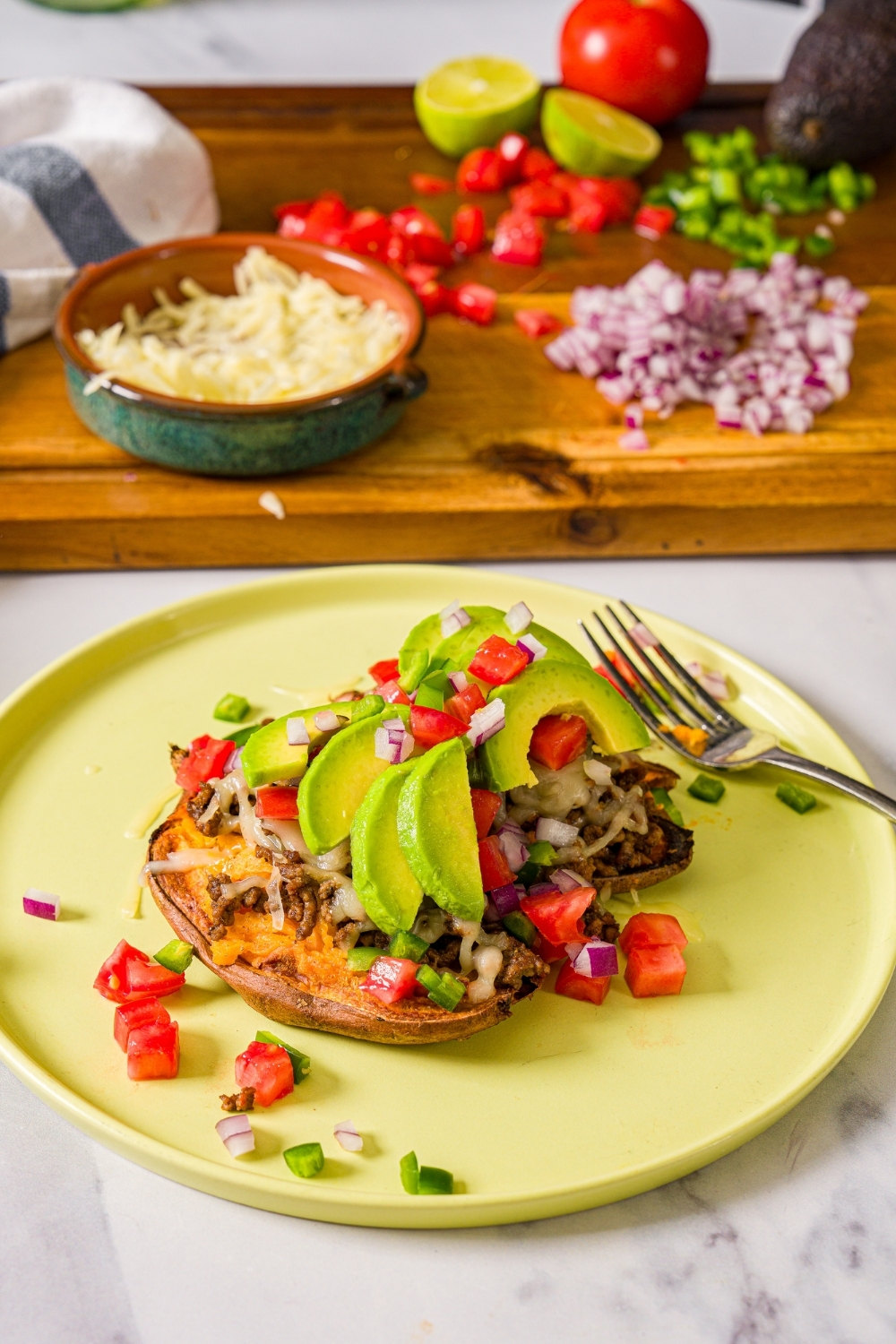 A green plate with an open baked sweet potato topped with seasoned taco meat, melted cheese, sliced avocado, and chopped tomatoes and red onion served with a halved lime. The plate is on a marble counter with a board of toppings.