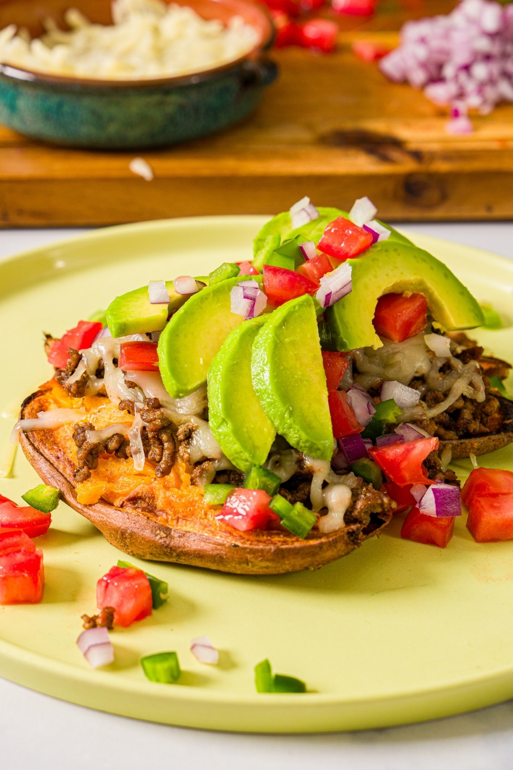 A green plate with an open baked sweet potato topped with seasoned taco meat, melted cheese, sliced avocado, and chopped tomatoes and red onion served with a halved lime. The plate is on a marble counter.