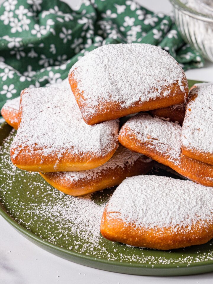 A green plate with a pile of beignets topped with honey and powdered sugar. The plate is on a marble counter with a green floral napkin.