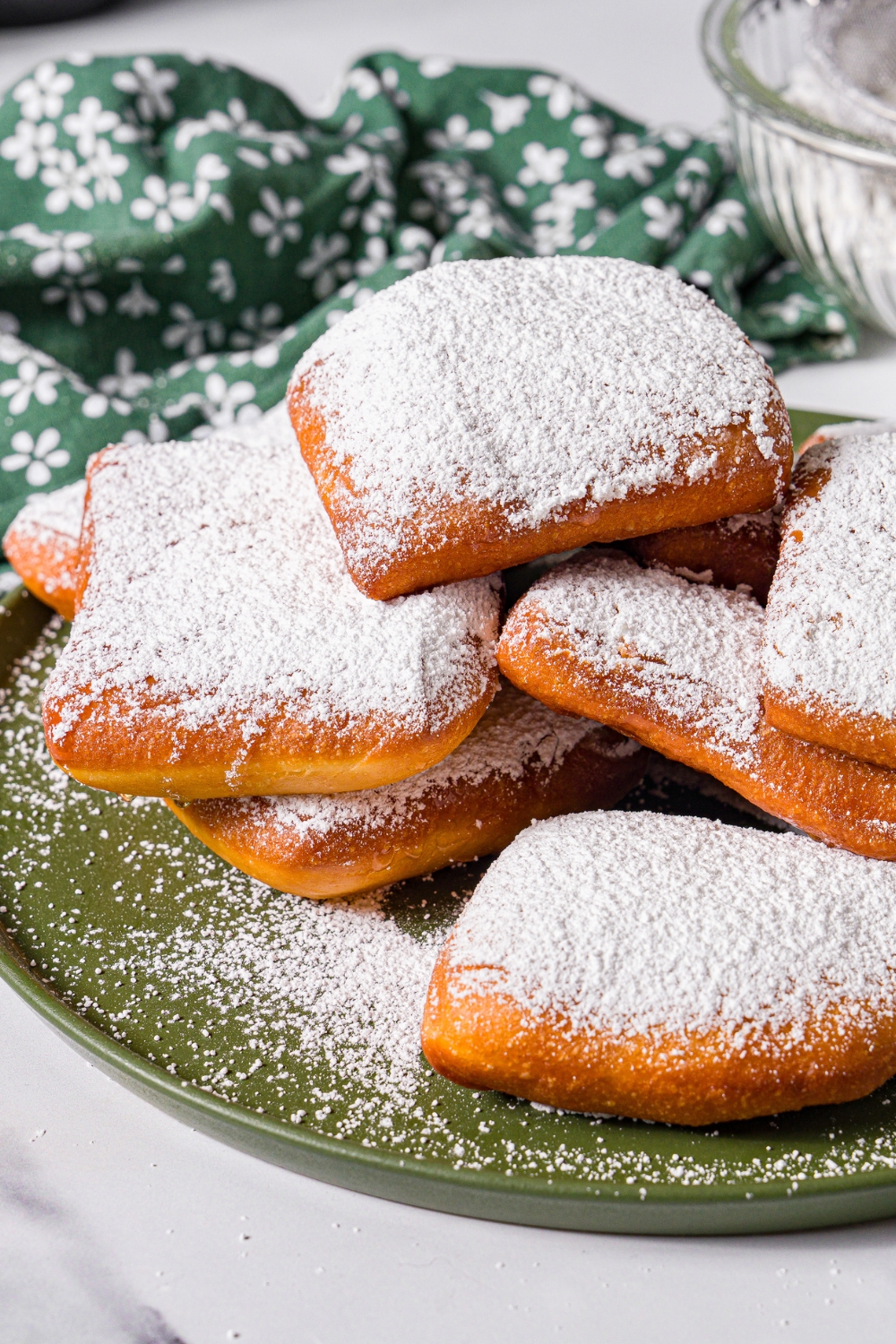 A green plate with a pile of beignets topped with honey and powdered sugar. The plate is on a marble counter with a green floral napkin.