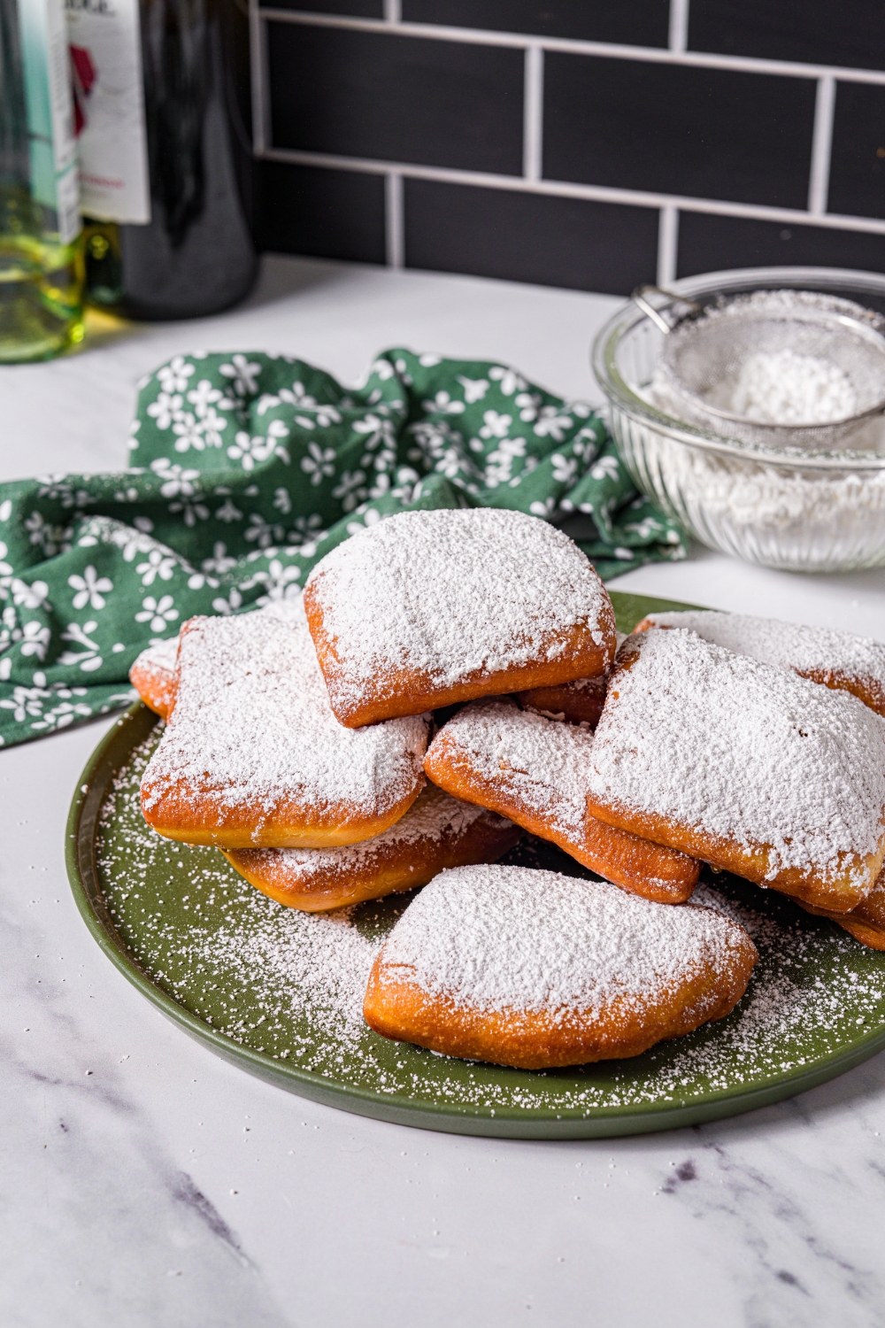 A green plate with a pile of beignets topped with honey and powdered sugar. The plate is on a marble counter with a green floral napkin.