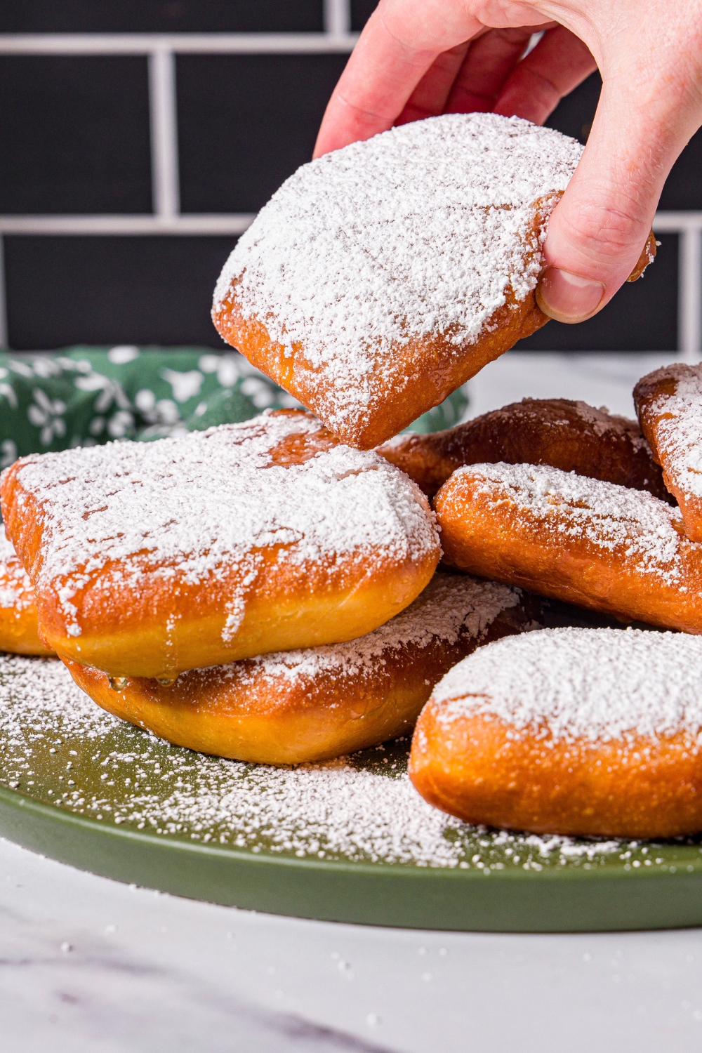 A green plate with a pile of beignets topped with honey and powdered sugar. A hand is taking a beignet from the plate. The plate is on a marble counter with a green floral napkin.