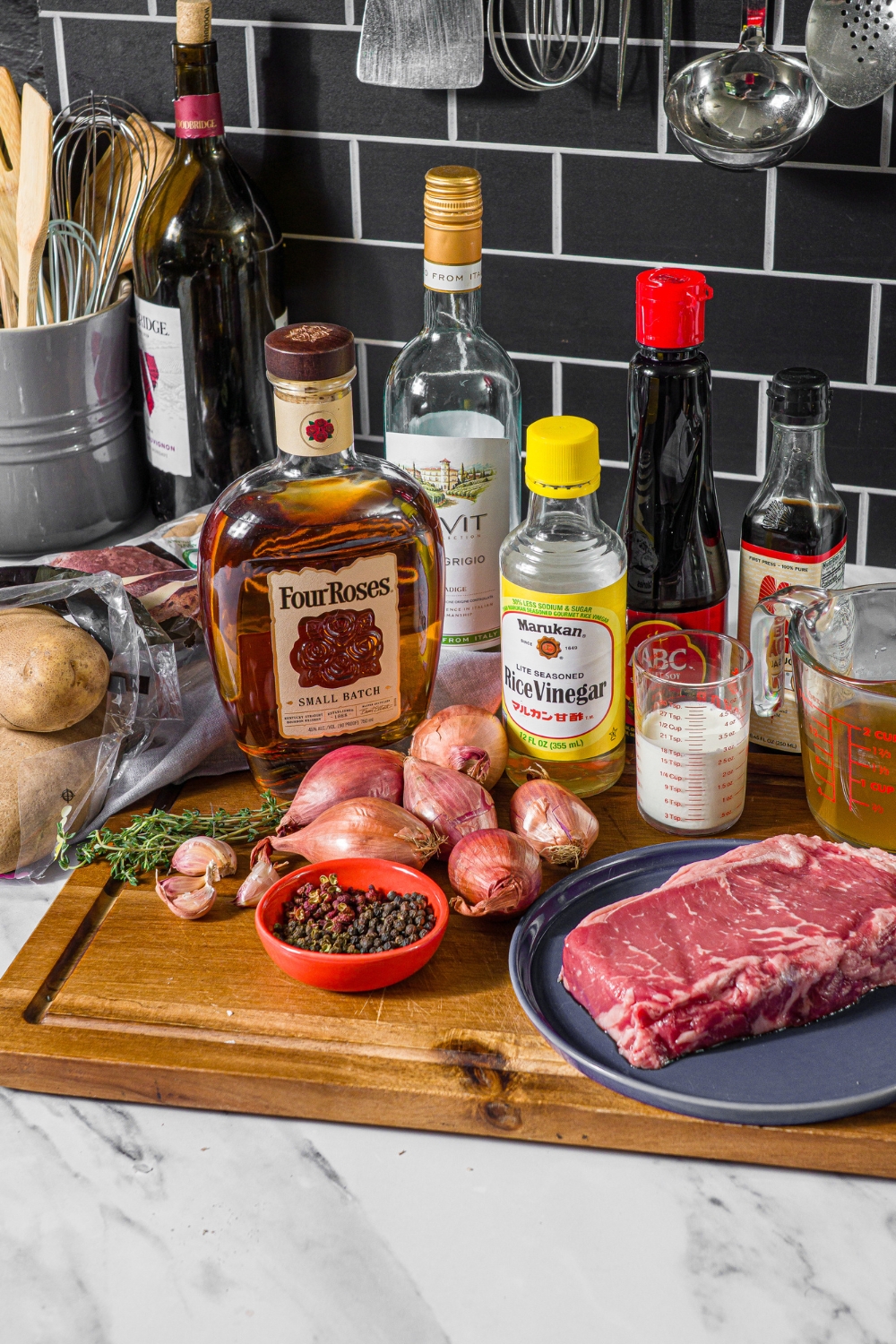 A wooden board with ingredients to make steak frites including steak, potatoes, rice wine vinegar, shallots, peppercorns, chicken stock, white wine, cream, soy sauce, and more.