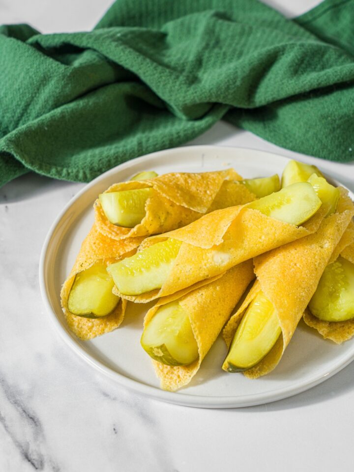 A white plate with a pile of chickles with pickles wrapped in cheese. The plate is on a marble counter with a green cloth napkin.