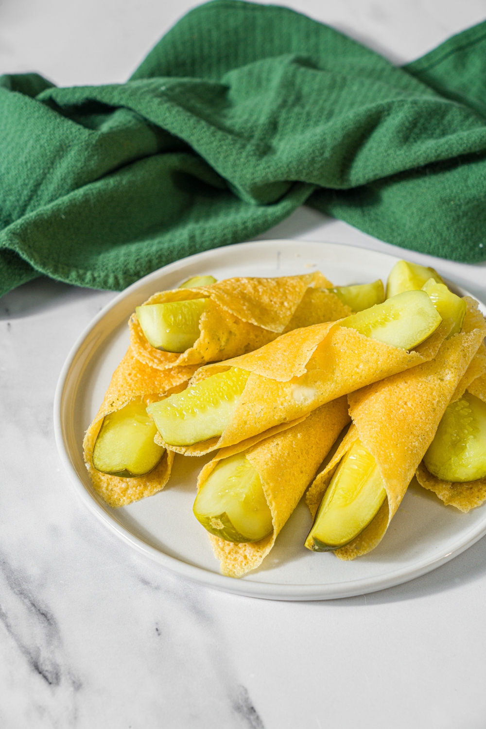 A white plate with a pile of chickles with pickles wrapped in cheese. The plate is on a marble counter with a green cloth napkin.