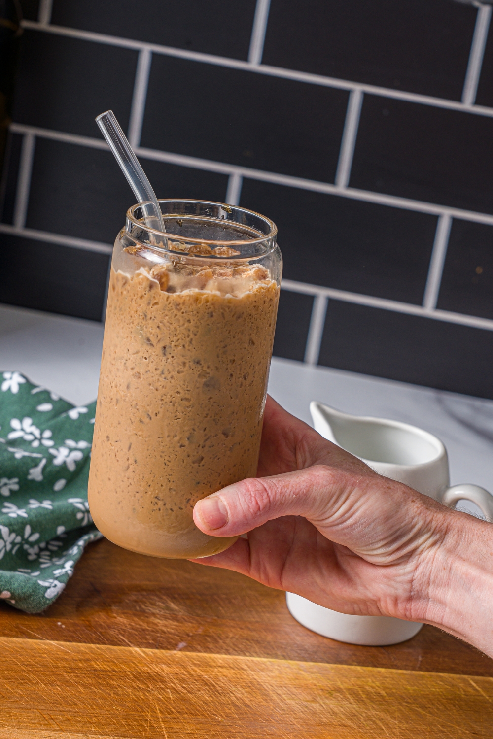 A hand holding a coffee slushie with a straw in the glass. There is a wooden board with a carafe of milk blurred in the background.