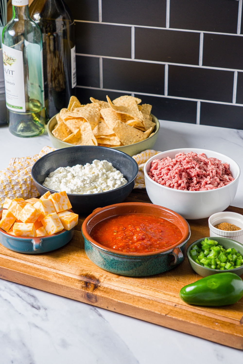 A wooden board with several bowls containing ingredients to make divorced dad dip including ground beef, salsa, cottage cheese, jalapeño, cheese cubes, and more.