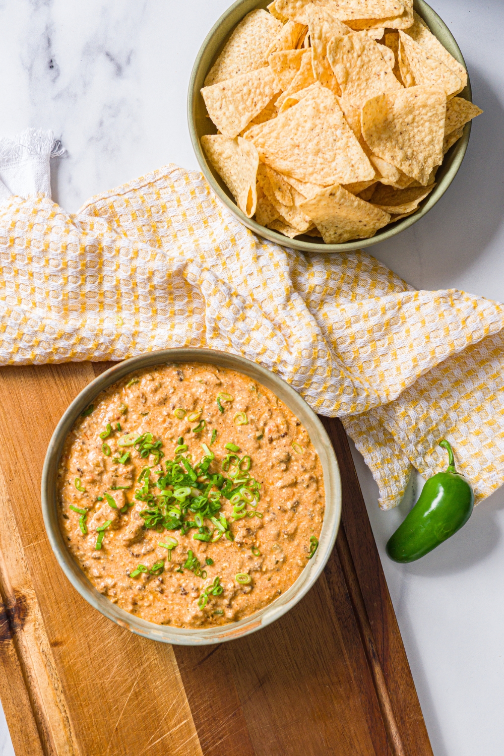 A bowl of divorced dad dip garnished with sliced green onion on a wooden board with a jalapeño. The board is on a white counter with a bowl of tortilla chips.