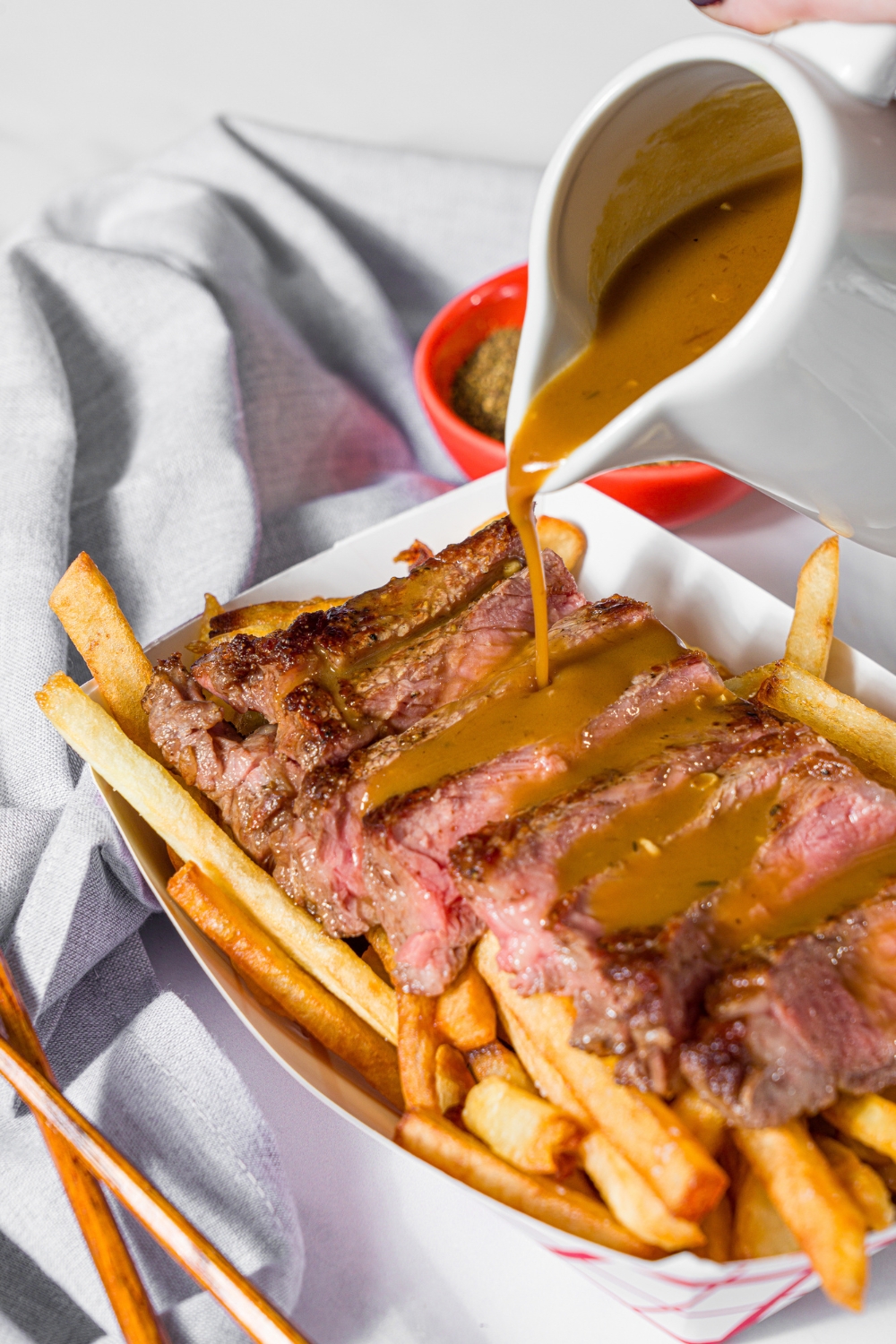 A creamer of steak frites sauce being poured over a fry basket with steak frites. The fry basket is on a marble counter.