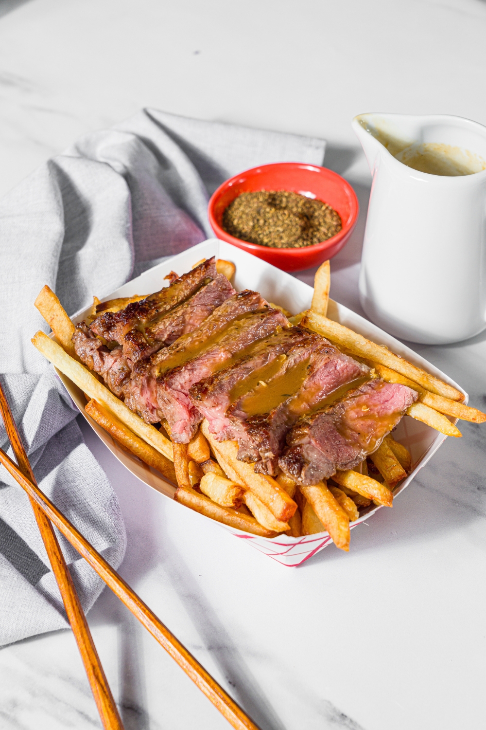 A fry basket with french fries topped with sliced steak and topped with steak frites sauce. The basket is on a marble counter with a bowl of seasonings.