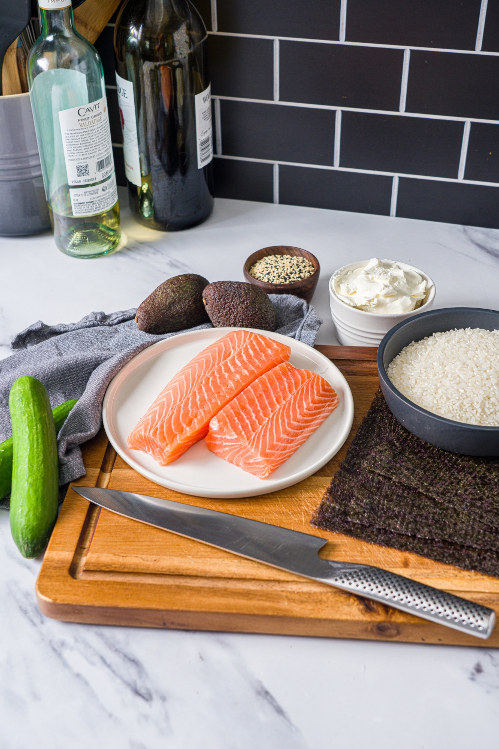 A wooden board with ingredients to make sushi cake including salmon, rice, nori sheets, avocado, cream cheese, avocado, and cucumber.