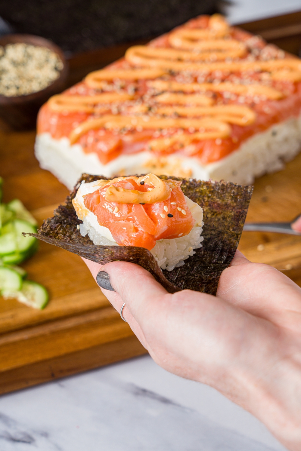 A hand holding a nori sheet topped with sushi cake with spicy mayo and sesame seeds. There is a wooden board with sushi cake blurred in the background.