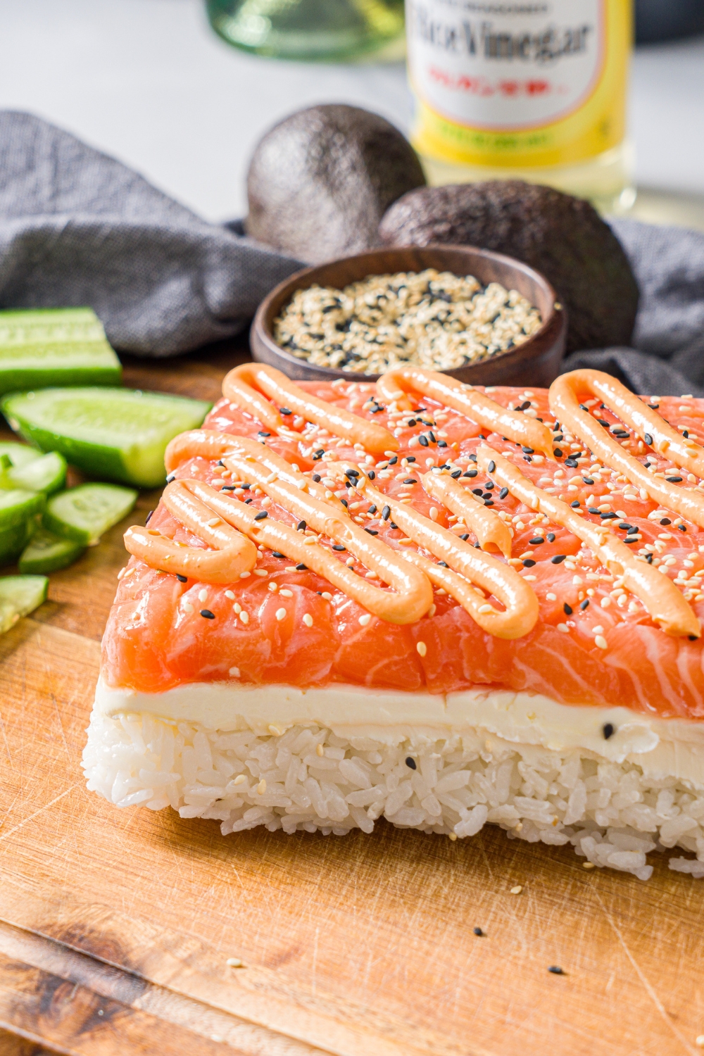 A wooden board with a whole salmon sushi cake topped with sesame seeds and spicy mayo. The board is on a marble counter with sliced cucumber and a bowl of sesame seeds.