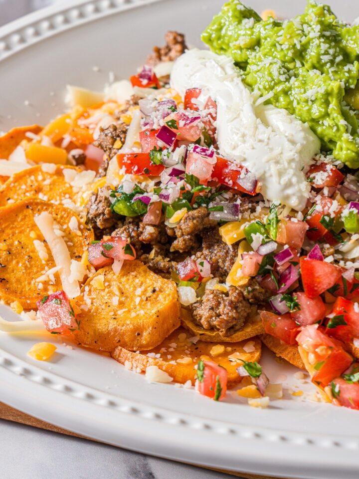 A white platter with sweet potato nachos including sweet potato medallions topped with seasoned ground beef, pico de gallo, cheese, sour cream, and guacamole.