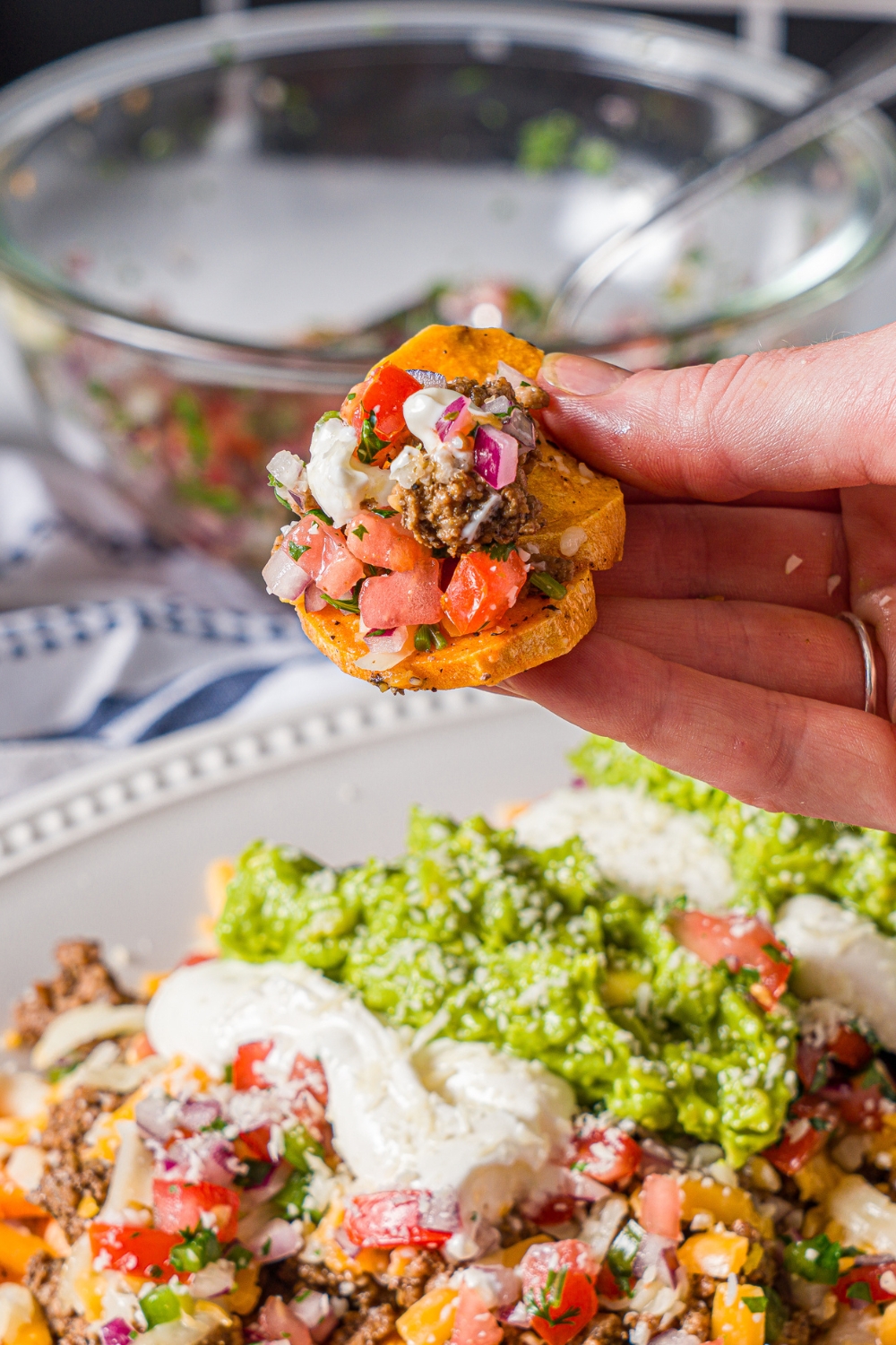 A hand holding a sweet potato nacho topepd with seasoned ground beef and pico de gallo. There is a platter of nachos blurred in the background.