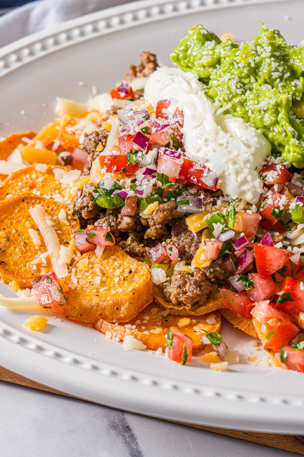A white platter with sweet potato nachos including sweet potato medallions topped with seasoned ground beef, pico de gallo, cheese, sour cream, and guacamole.