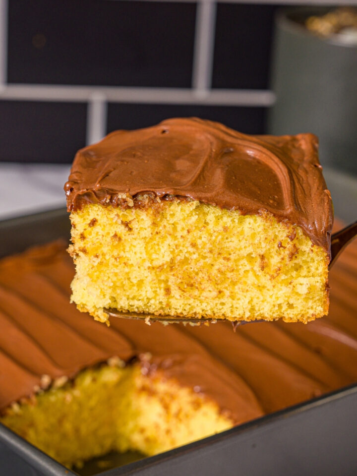 A slice of yellow cake with chocolate frosting being removed from a baking pan of cake.
