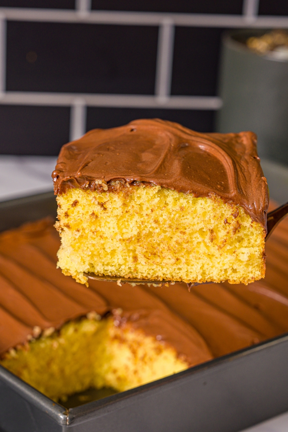 A slice of yellow cake with chocolate frosting being removed from a baking pan of cake.