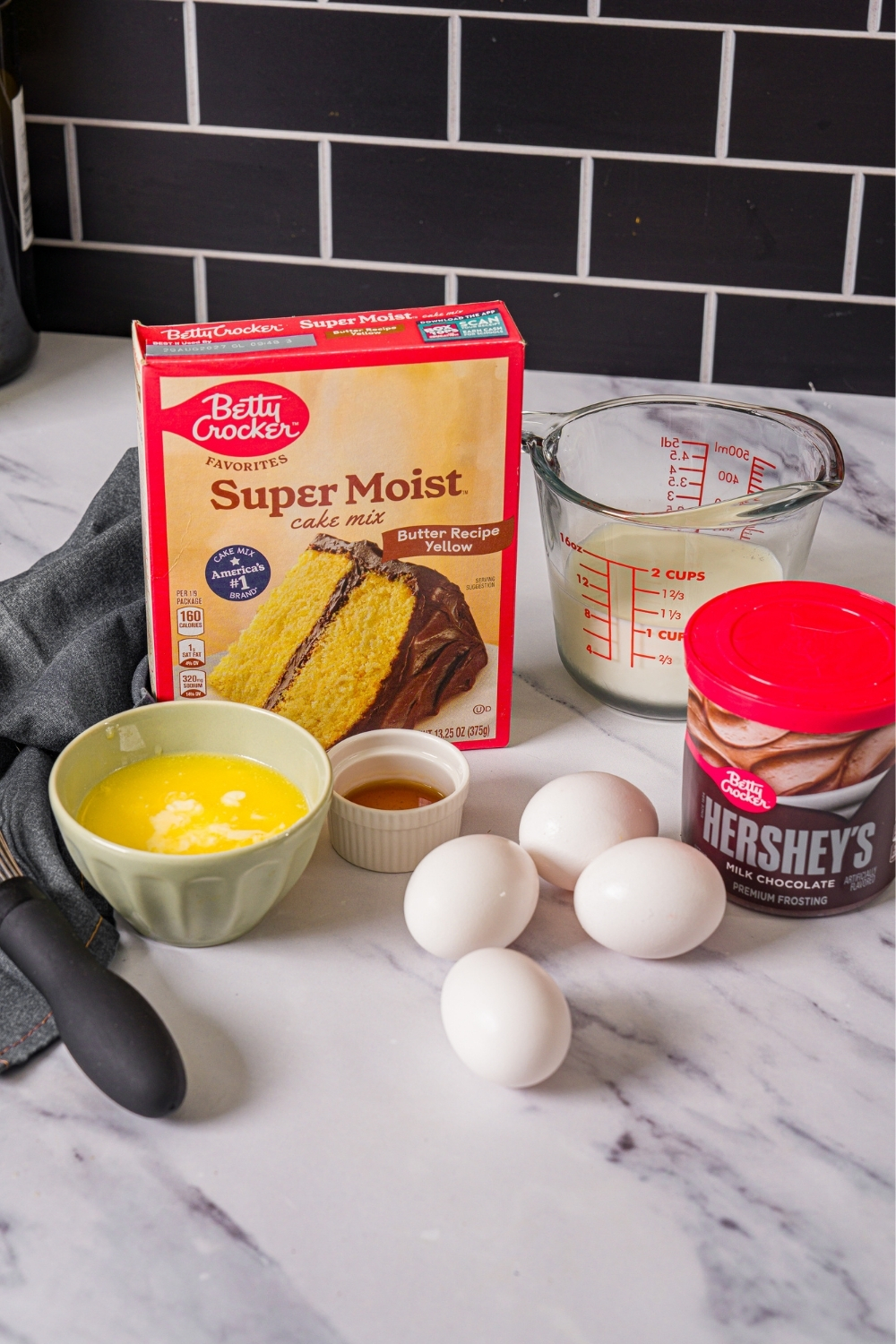 A marble counter with ingredients to make yellow cake including yellow cake mix, eggs, butter, milk, vanilla, and chocolate frosting.