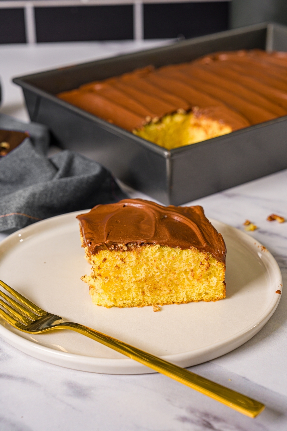 A white plate with a slice of yellow cake with chocolate frosting with a fork on the plate. The plate is on a marble counter with a baking pan of cake.