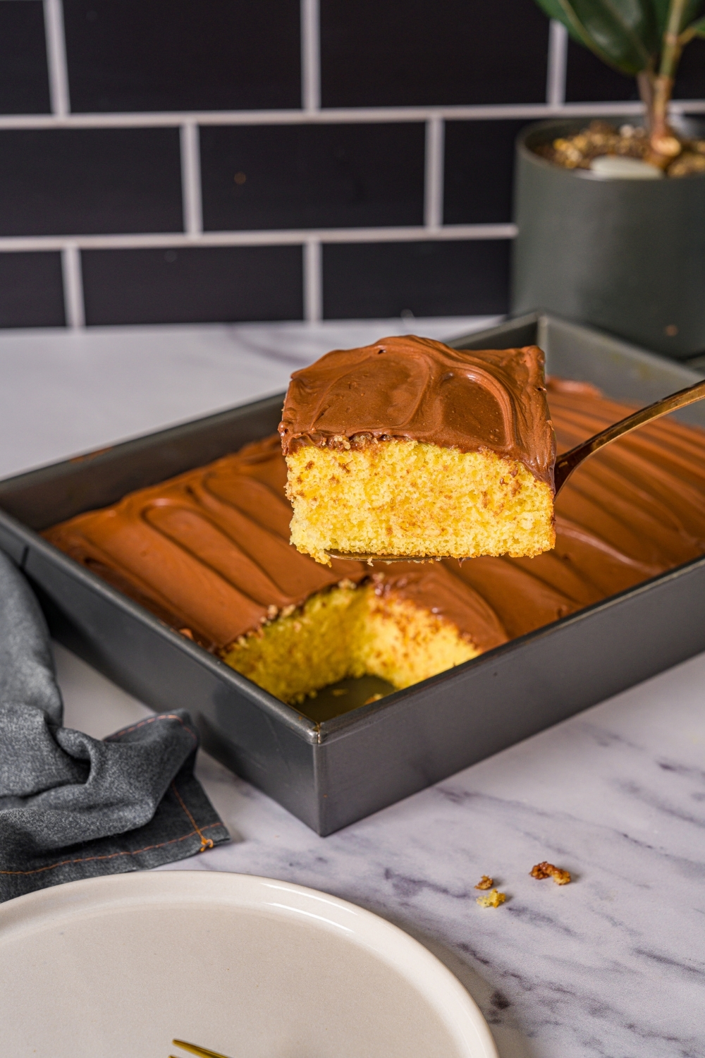 A slice of yellow cake with chocolate frosting being removed from a baking pan of cake. The pan is on a marble counter with a white plate.