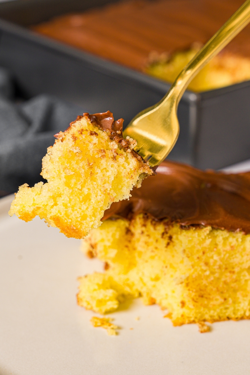 A close up of a fork with a bite of yellow cake with chocolate frosting. There is a plate with cake on a marble counter blurred in the background.