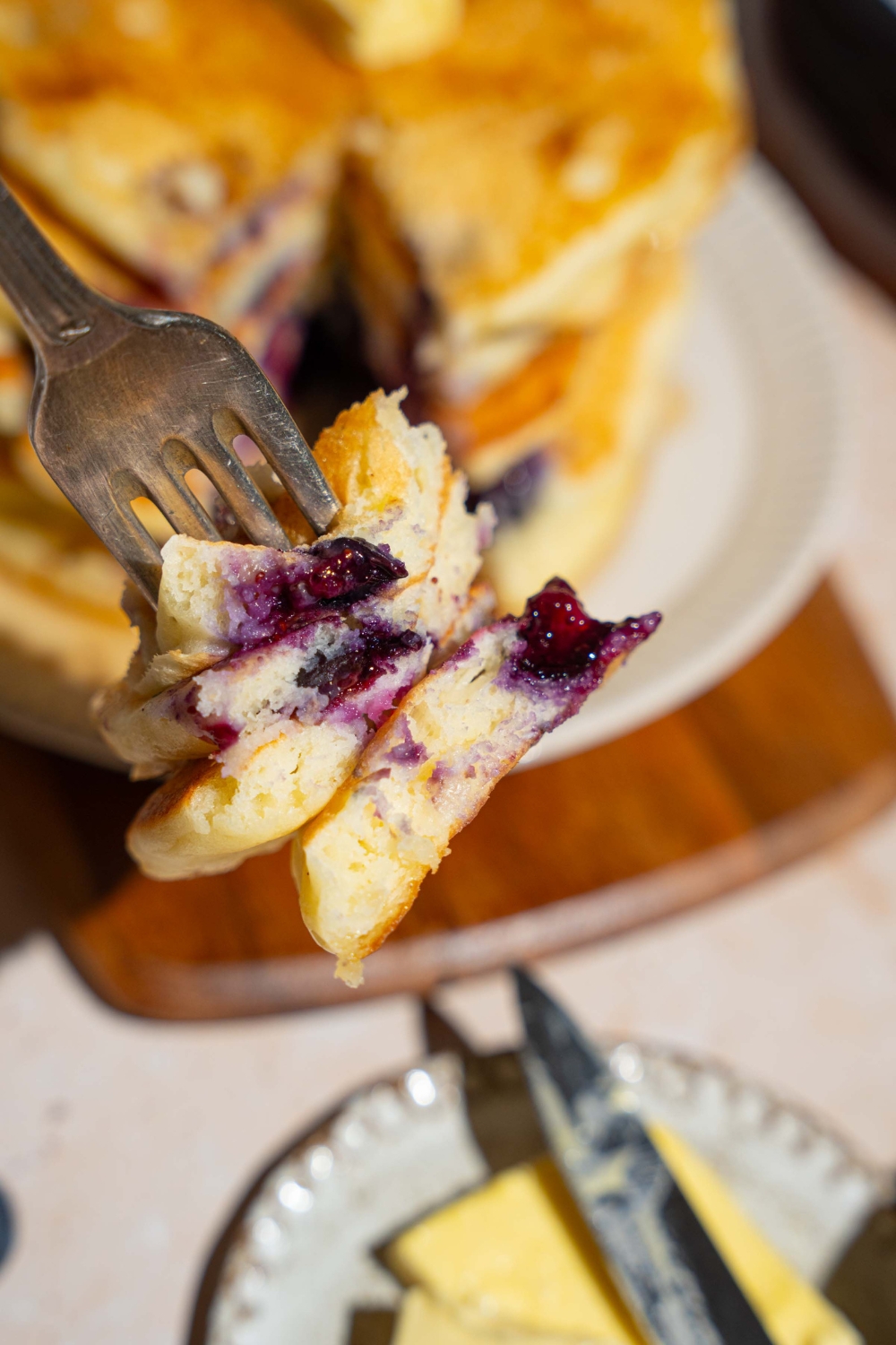 A close up of a fork with a bite of buttermilk blueberry pancakes. There is a plate of pancakes on a wooden board blurred in the background.