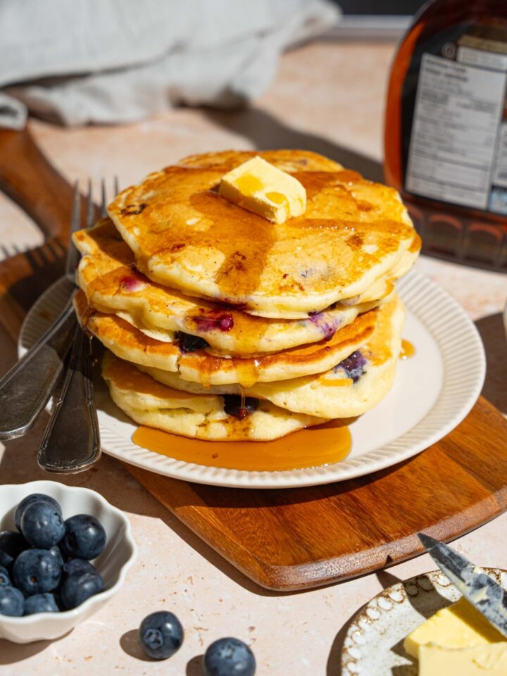 A white plate with a stack of buttermilk blueberry pancakes topped with butter and drizzled with syrup. There is a fork and knife on the plate. The plate is on a wooden board on a marble counter with a bowl of blueberries and syrup.