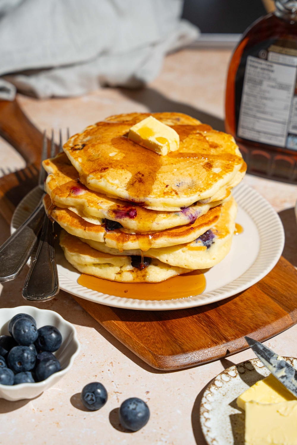 A white plate with a stack of buttermilk blueberry pancakes topped with butter and drizzled with syrup. There is a fork and knife on the plate. The plate is on a wooden board on a marble counter with a bowl of blueberries and syrup.