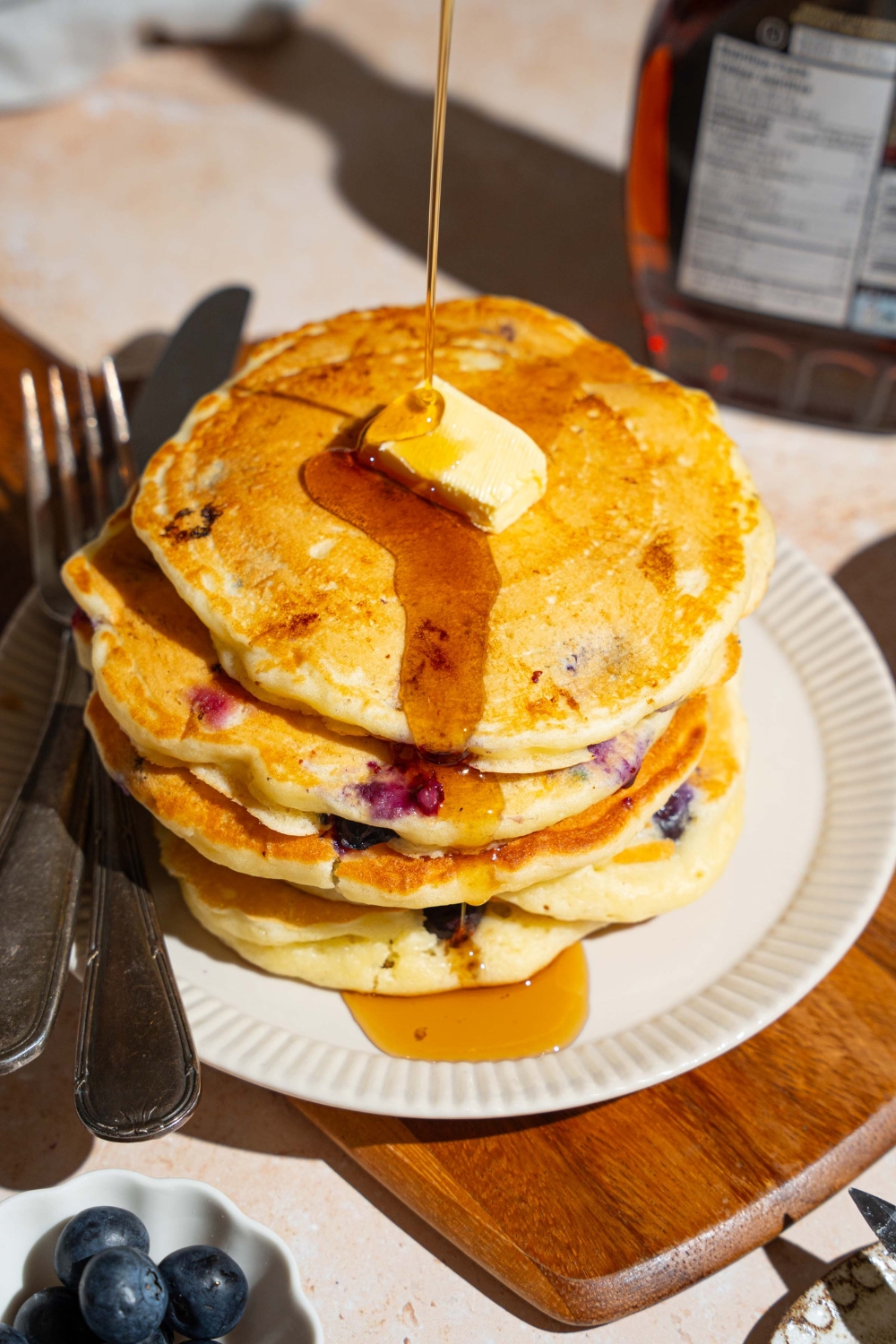A white plate with a stack of buttermilk blueberry pancakes topped with butter with syrup being poured over the stack. There is a fork and knife on the plate. The plate is on a wooden board on a marble counter with a bowl of blueberries and syrup.