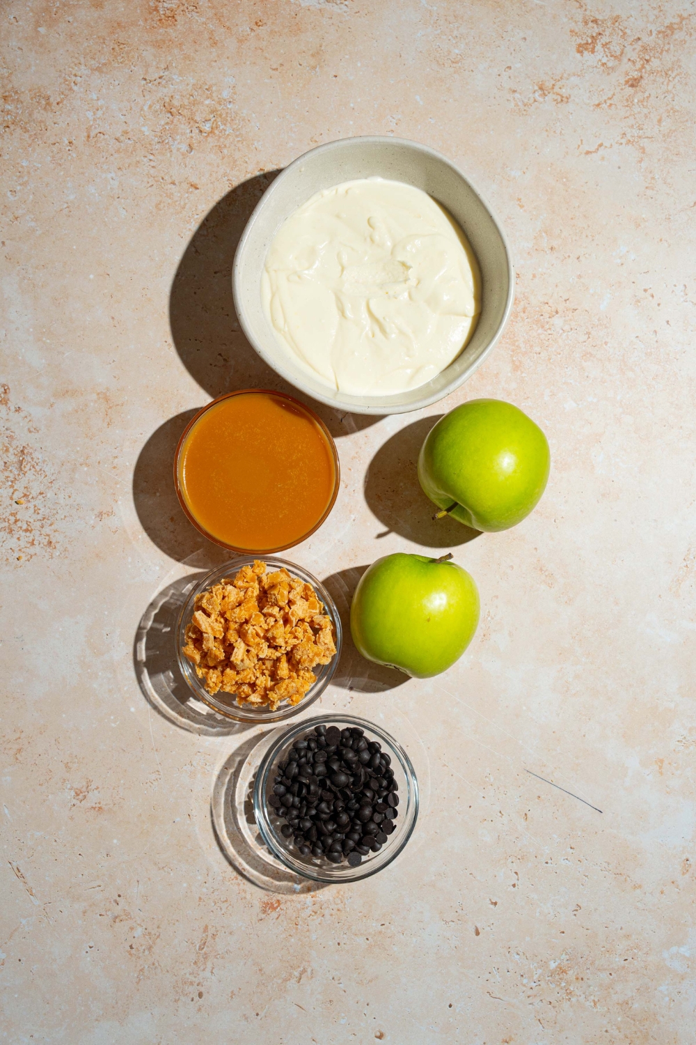 A tan counter with ingredients to make caramel apple cheesecake dip including cheesecake filling, apples, caramel, toffee bits, and chocolate chips.