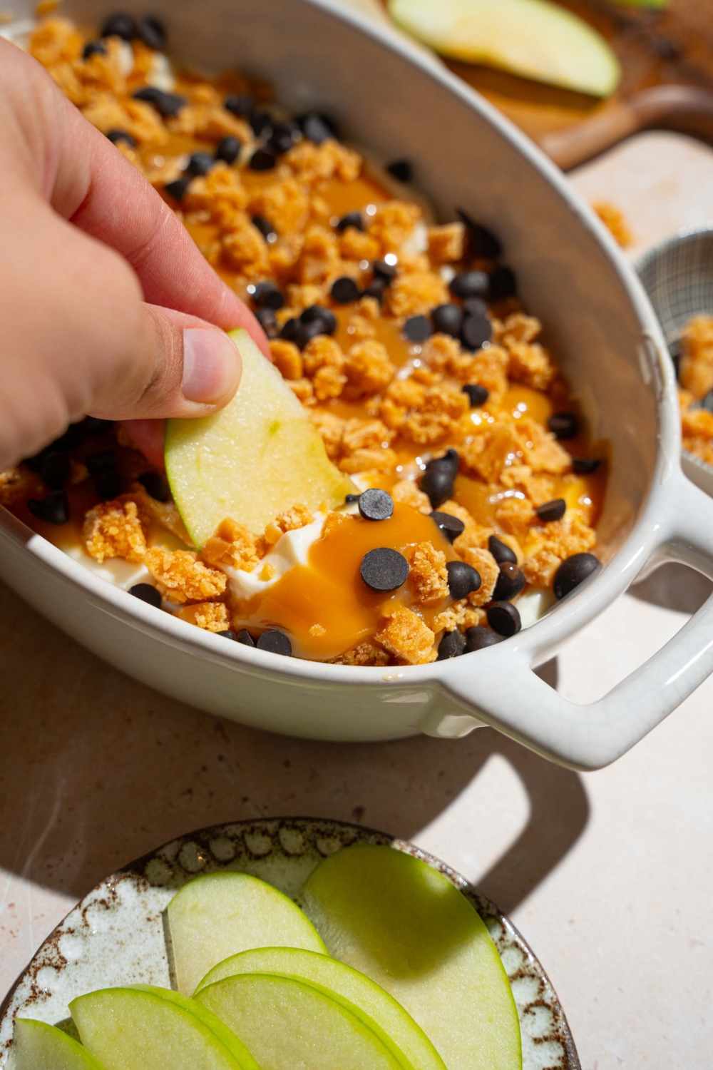 An oval platter with caramel apple cheesecake dip topped with toffee bits and chocolate chips. A hand is dipping an apple slice into the dip. The platter is on a tan counter with a plate of sliced apples.