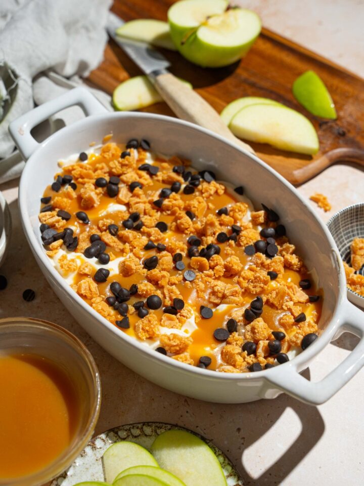 An oval platter with caramel apple cheesecake dip topped with toffee bits and chocolate chips. The platter is on a tan counter with a plate of sliced apples and bowl of caramel sauce.