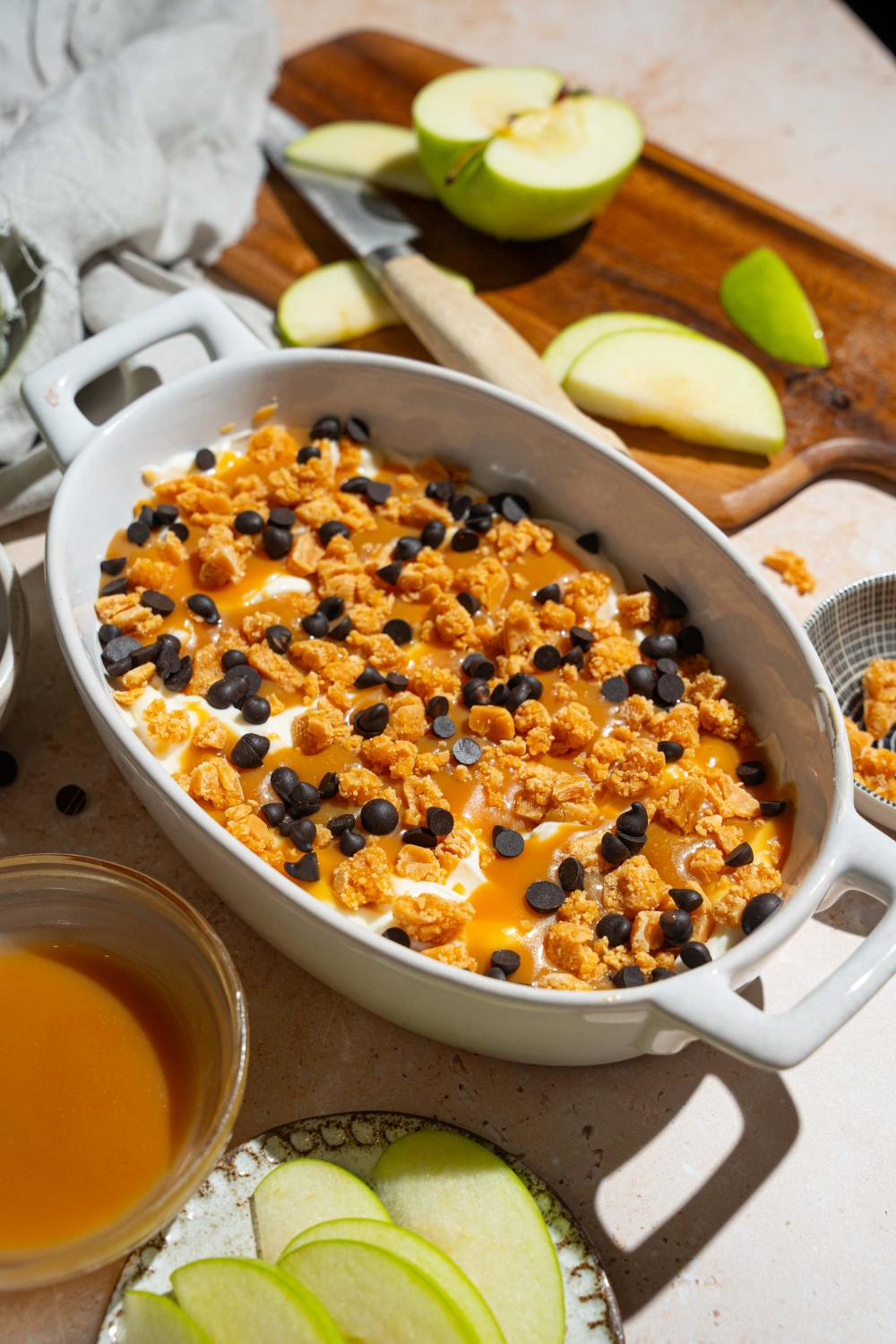An oval platter with caramel apple cheesecake dip topped with toffee bits and chocolate chips. The platter is on a tan counter with a plate of sliced apples and bowl of caramel sauce.