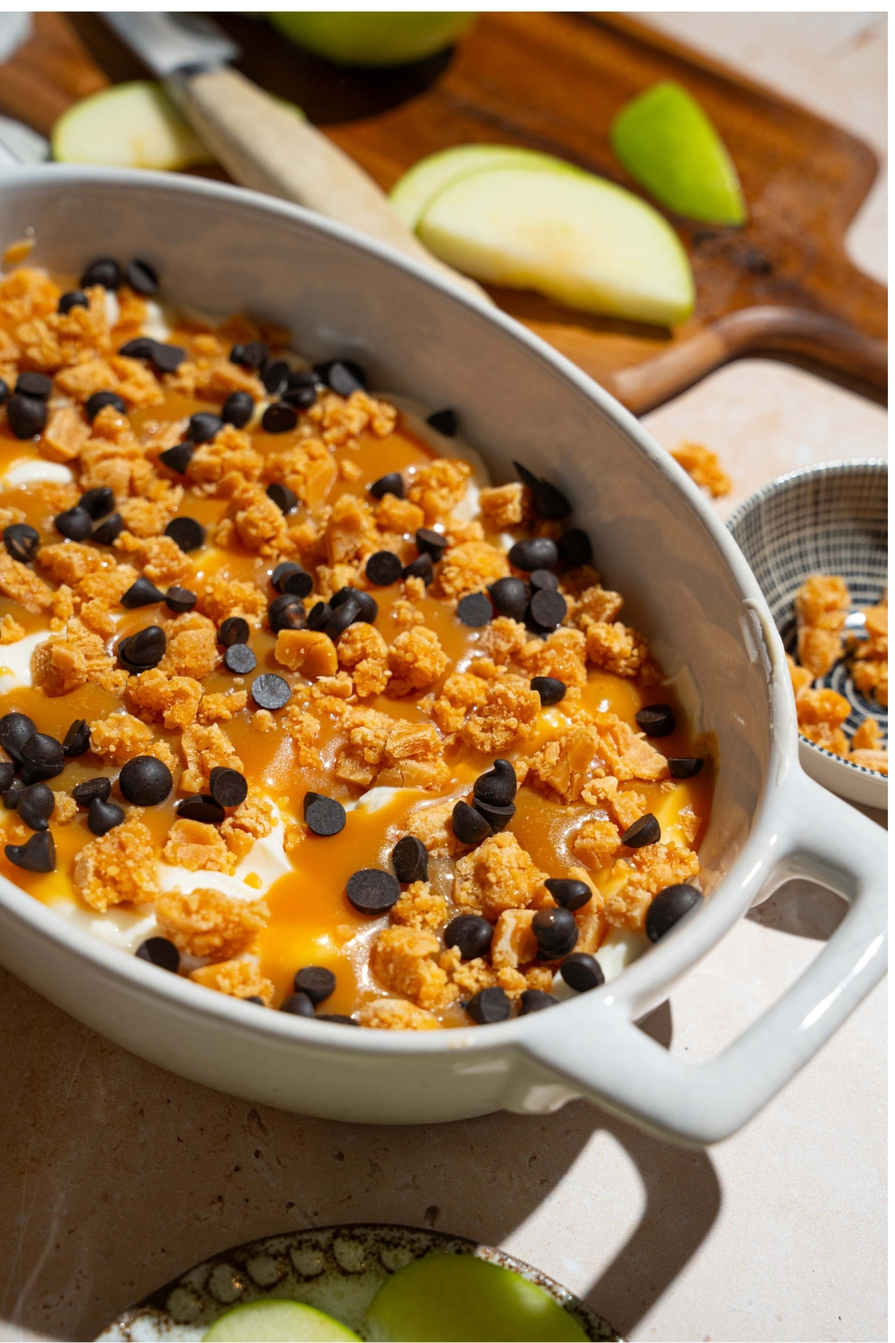 An oval platter with caramel apple cheesecake dip topped with toffee bits and chocolate chips. The platter is on a tan counter with a board of sliced apples.