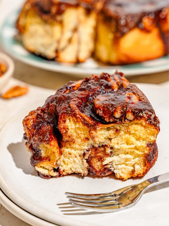 A caramel pecan sticky bun on a white plate that is in front of a tray that has the rest of the pecan sticky buns on it. Both are on a white counter top.