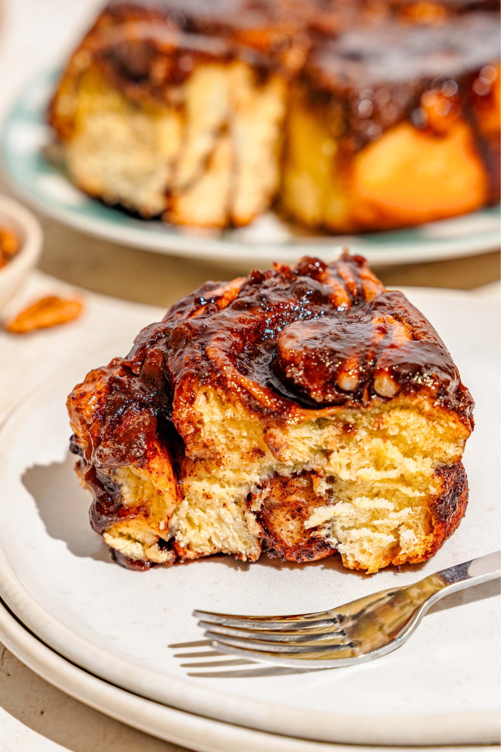 A caramel pecan sticky bun on a white plate that is in front of a tray that has the rest of the pecan sticky buns on it. Both are on a white counter top.