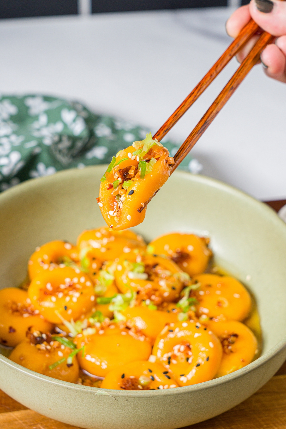 A green bowl with carrot button noodles tossed in a chili crisp sauce and garnished with sliced green onions. A pair of chopsticks is taking a bite from the bowl. The bowl is on a wooden board on a marble counter with a green floral napkin.