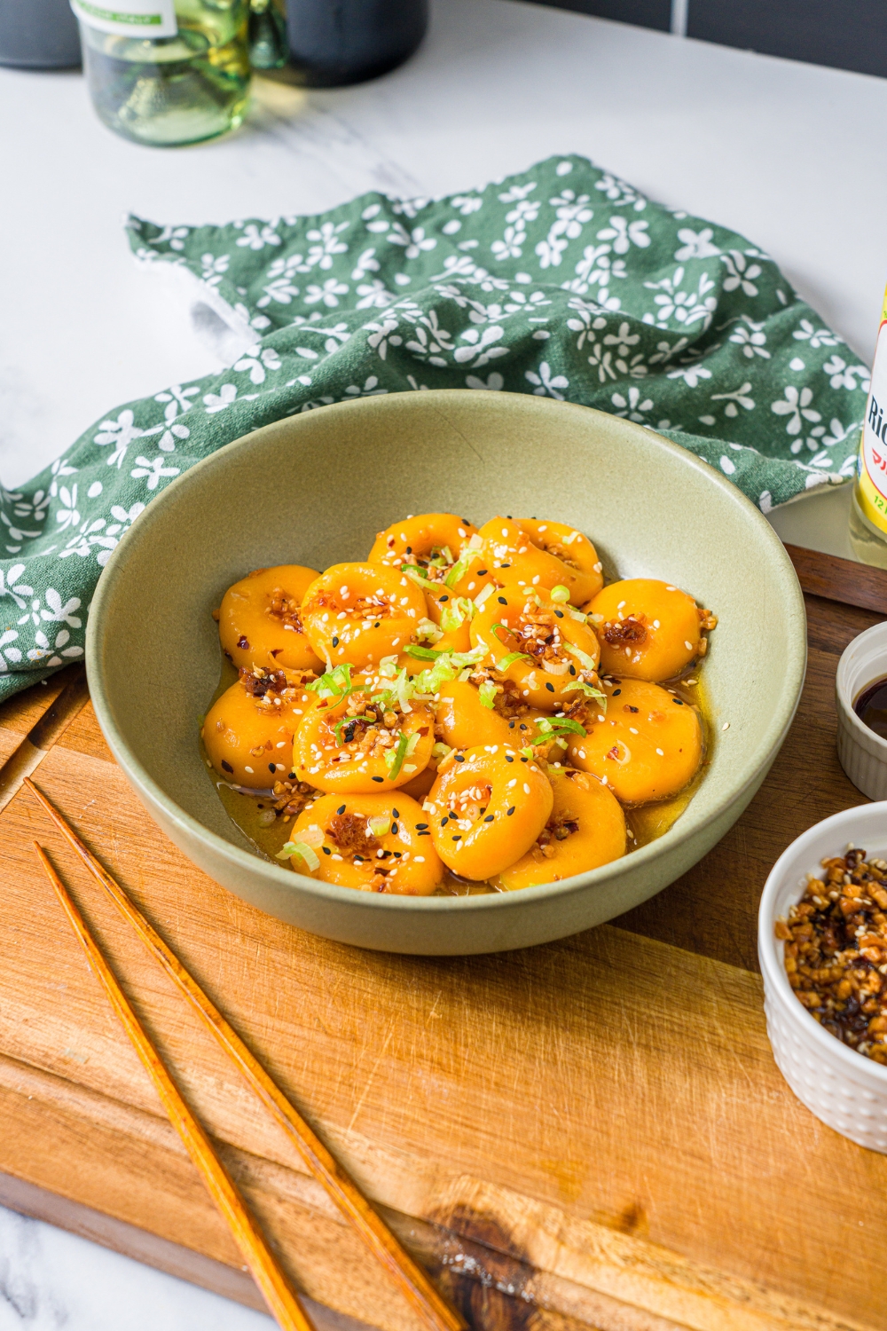 A green bowl with carrot button noodles tossed in a chili crisp sauce and garnished with sliced green onions. The bowl is on a wooden board on a marble counter with a green floral napkin.