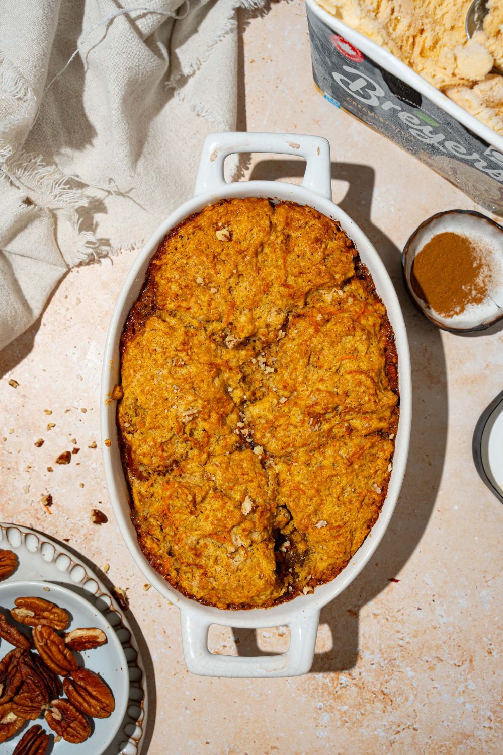 An oval baking dish with carrot cake cobbler with pecan pie filling. The dish is on a tan counter with a white cloth napkin and bowl of pecans.