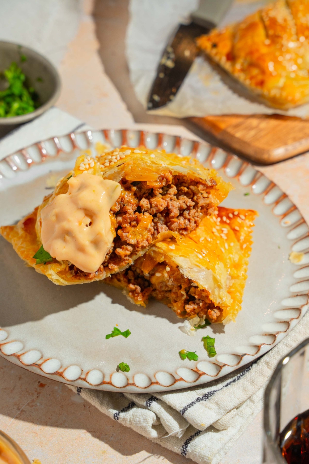 A white plate with a sliced cheeseburger hot pocket garnished with fresh parsley and dipped in animal sauce. The plate is on a white striped napkin on a tan counter with a tray of hot pockets.