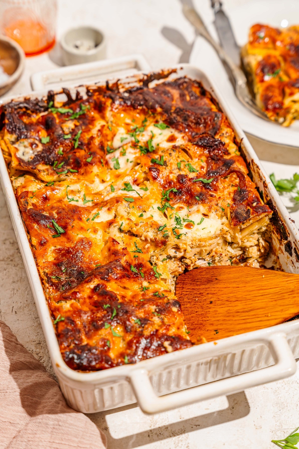 A baking dish with baked chicken alfredo lasagna garnished with fresh parsley. A wooden spoon has removed a slice from the dish. The dish is on a tan counter with a plate of lasagna.