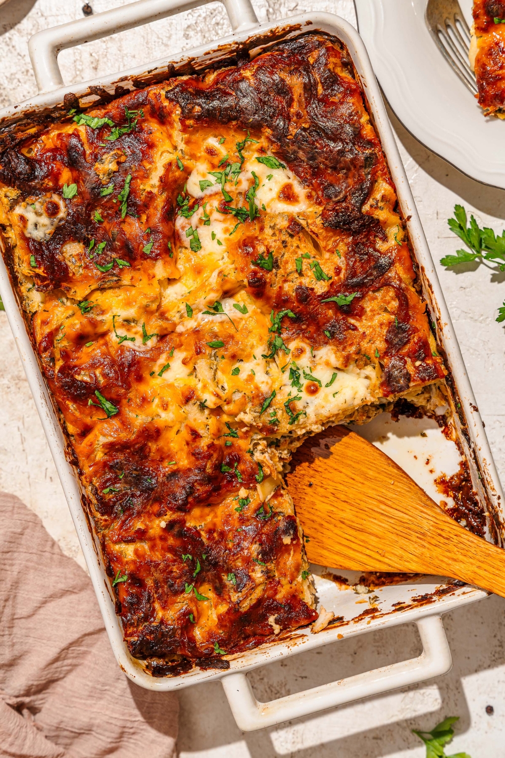 A baking dish with baked chicken alfredo lasagna garnished with fresh parsley. A wooden spoon has removed a slice from the dish. The dish is on a tan counter with a brown cloth napkin.