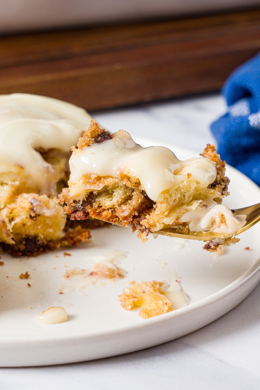 A fork with a piece of cookie dough cinnamon roll on it. Behind it is a white plate with the cinnamon roll on it.