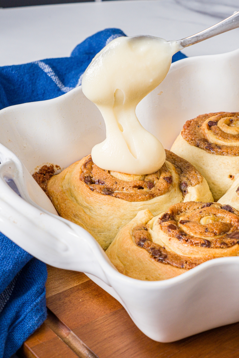 A spoon spread spreading frosting on top of the cookie dough cinnamon roll that is in a white tray with a few more cookie dough cinnamon rolls.