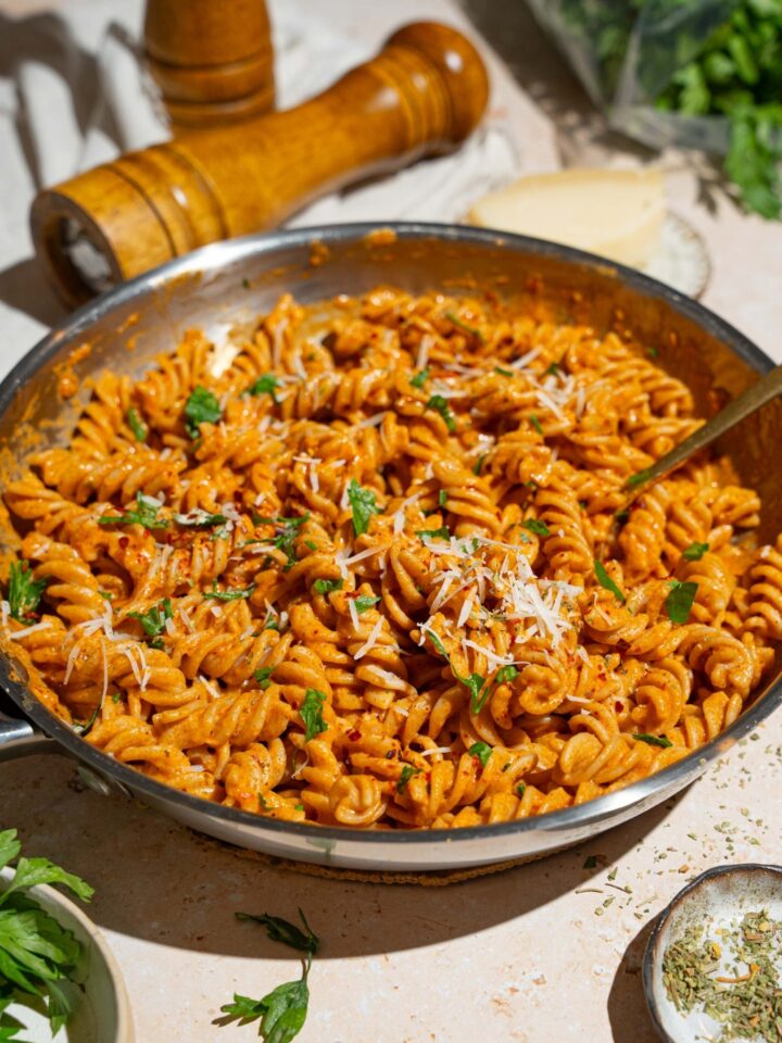 A skillet with fusilli pasta tossed in creamy tomato pasta sauce garnished with fresh basil and shredded cheese. There is a fork in the pasta. The skillet is on a tan counter with small bowls of garnishes and a pepper mill.