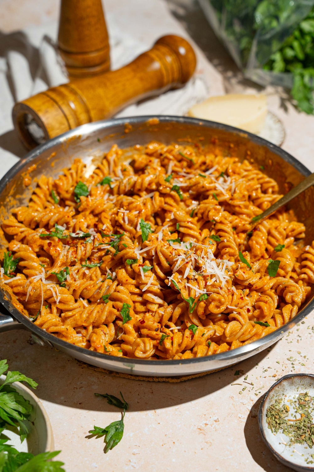 A skillet with fusilli pasta tossed in creamy tomato pasta sauce garnished with fresh basil and shredded cheese. There is a fork in the pasta. The skillet is on a tan counter with small bowls of garnishes and a pepper mill.